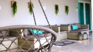 Una silla de mimbre con un asiento de cuerda, un sofá con un cojín azul verdoso, y plantas verdes en la pared.