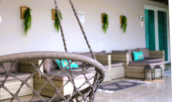 Ein Korbstuhl mit Netz aus Seil, ein Sofa mit einem türkisfarbenen Kissen und grüne Pflanzen an der Wand.