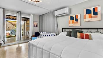 A modern hotel room with a large bed, a balcony with curtains, an air conditioning unit, and abstract wall art.
