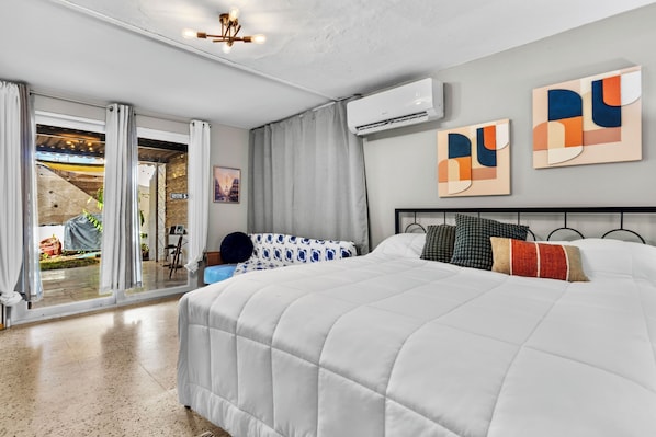 A modern hotel room with a large bed, a balcony with curtains, an air conditioning unit, and abstract wall art.