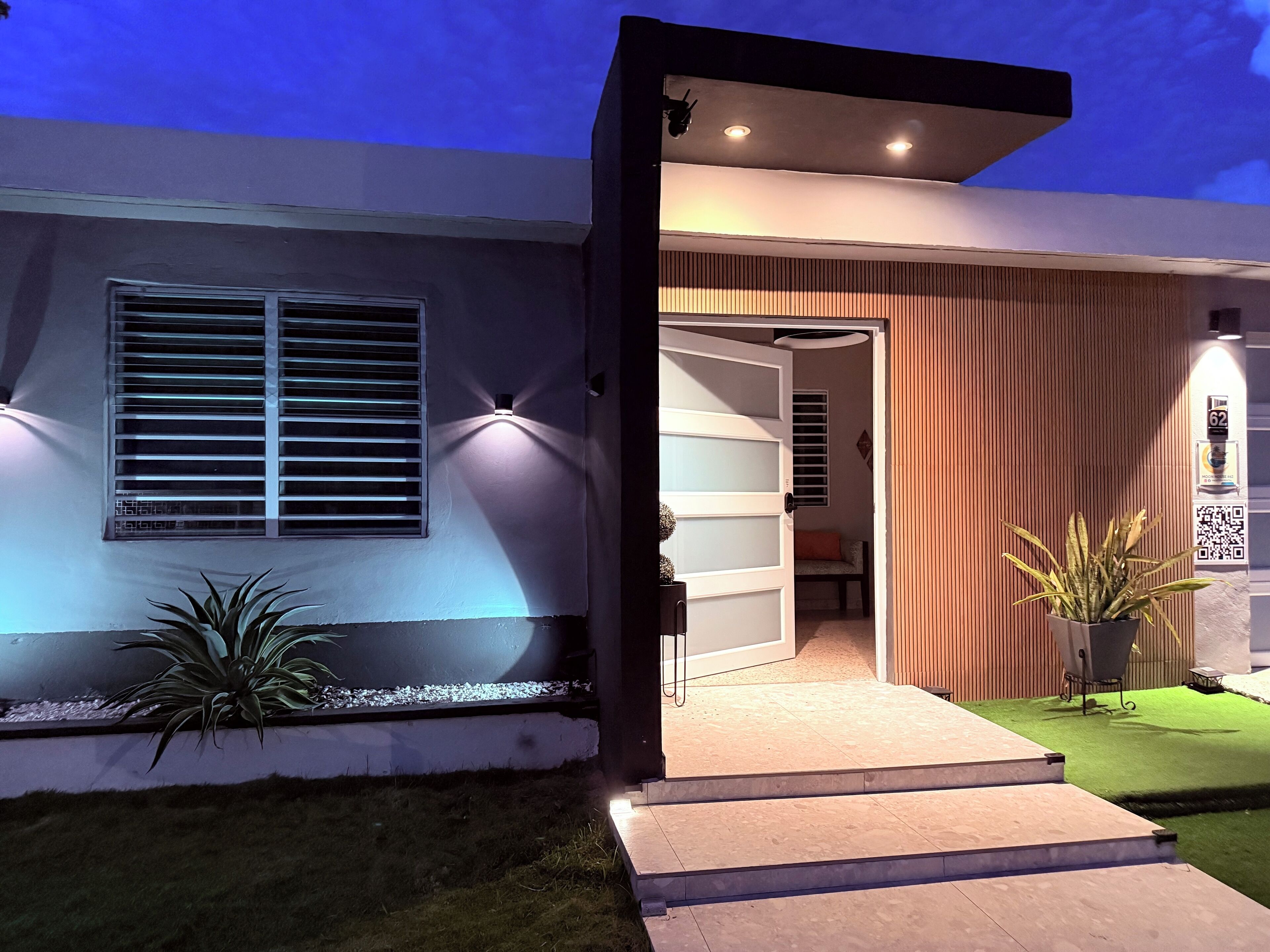 A modern house with a well-lit entrance, a visible door, and a small outdoor seating area.