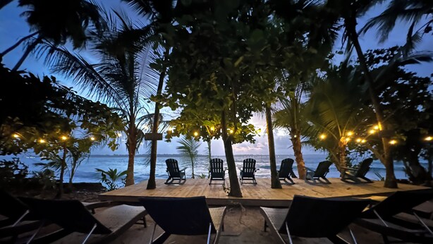 Un área de descanso junto a la playa con sillas y mesas, palmeras y vista al océano.