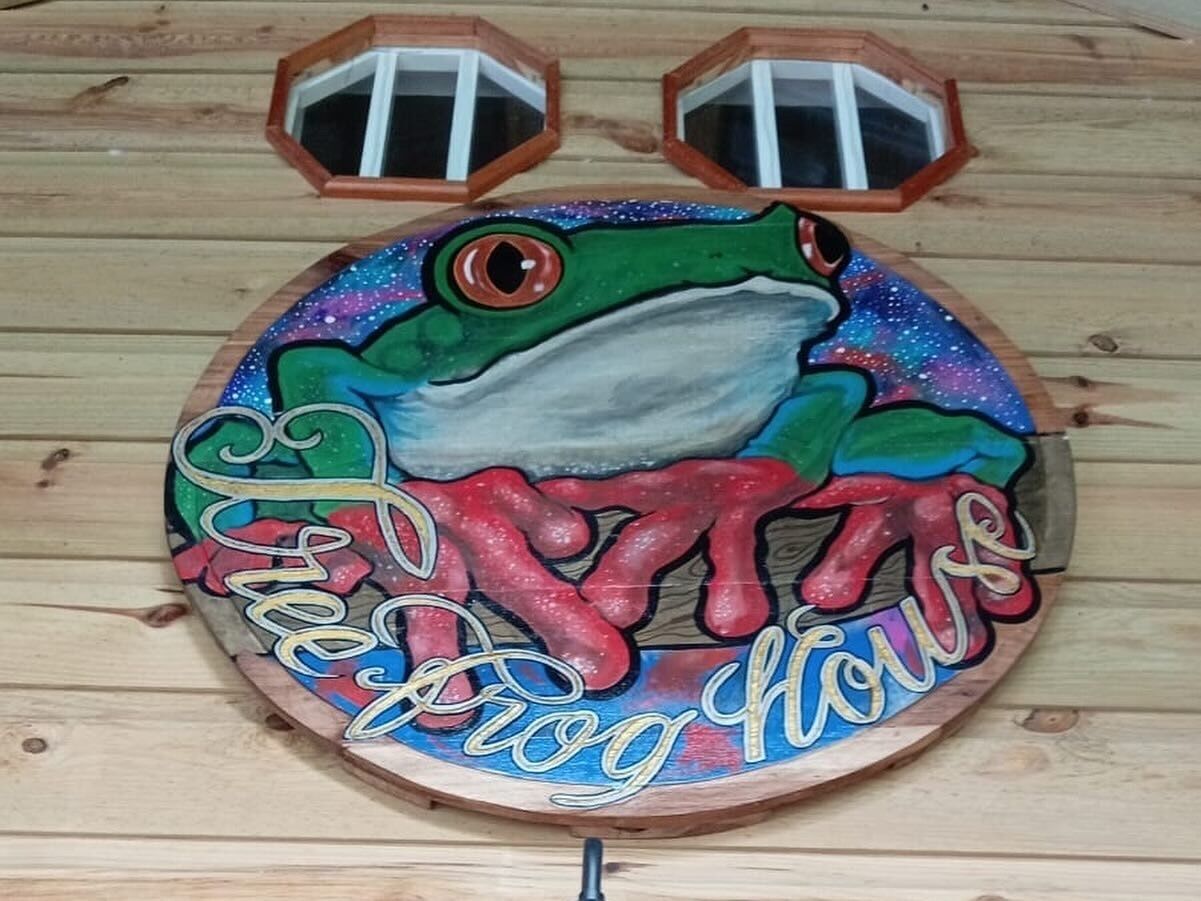 En rund träskylt med en groddesign och texten ”Frog House” på.