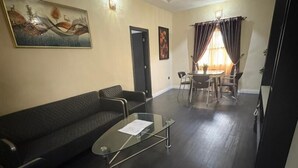 A living room with a dark-colored sofa, a glass coffee table, and a dining area with a table and chairs. - Cozy 1-BEDROOM WITH AC, WiFi, FULL kitchen, free parking, restaurant delivery (Satellite Town)