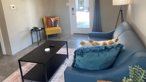 A modern living room with a black coffee table, a blue sofa with a decorative pillow, a yellow armchair with a colorful cushion, a floor lamp, and a TV mounted on the wall.
