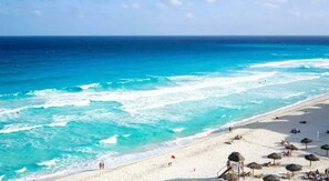 Una playa con agua turquesa cristalina, arena blanca y sombrillas de paja.