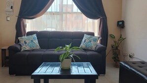 A living room with a dark brown sofa, two patterned pillows, a small table with a potted plant, and a window with curtains. - Alberca privada con cascada familia/amigos

 (Cancún)