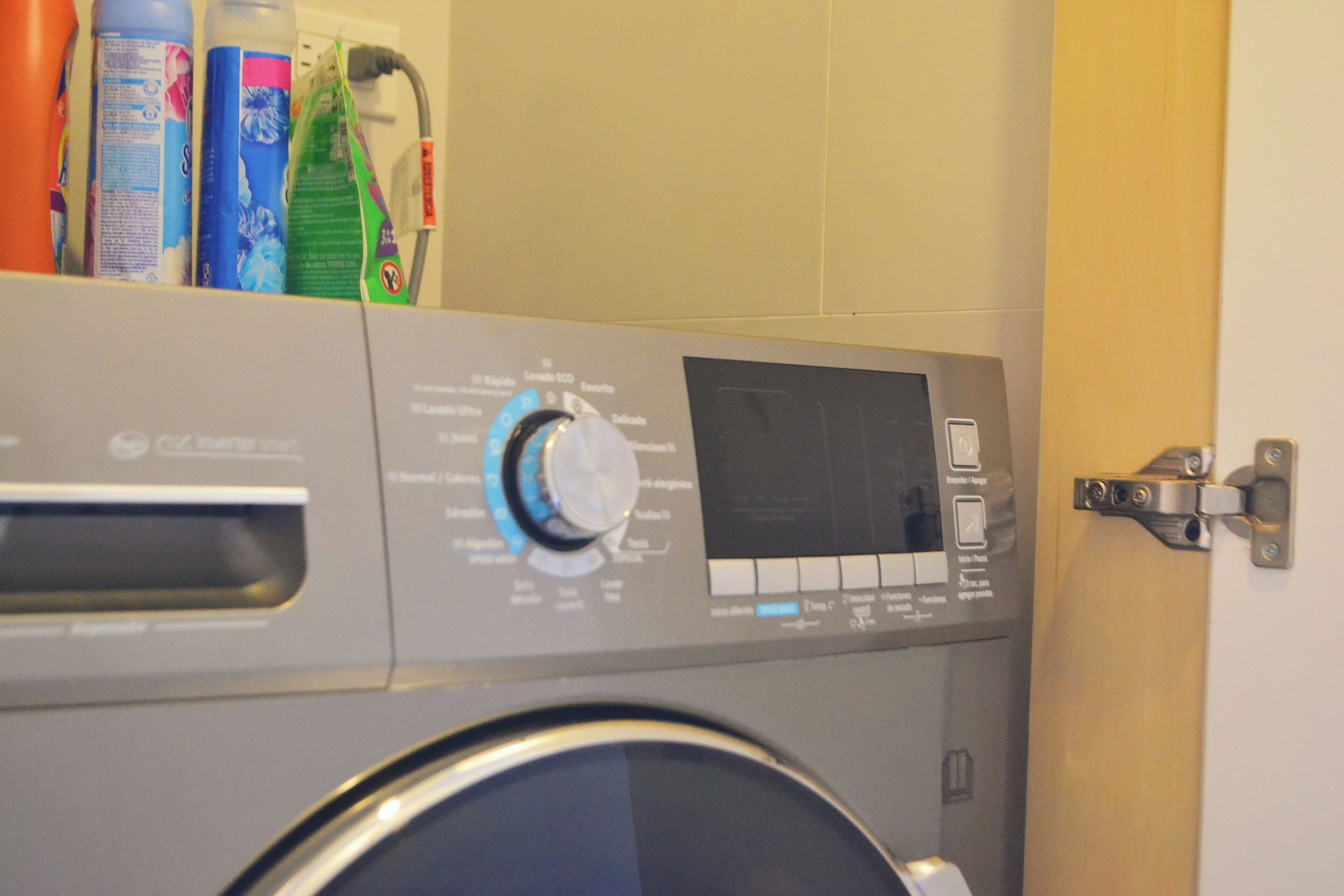 Un lave-linge à chargement frontal doté d’un panneau de commande, d’une porte et d’une étagère fixée au mur pour ranger les produits de nettoyage.