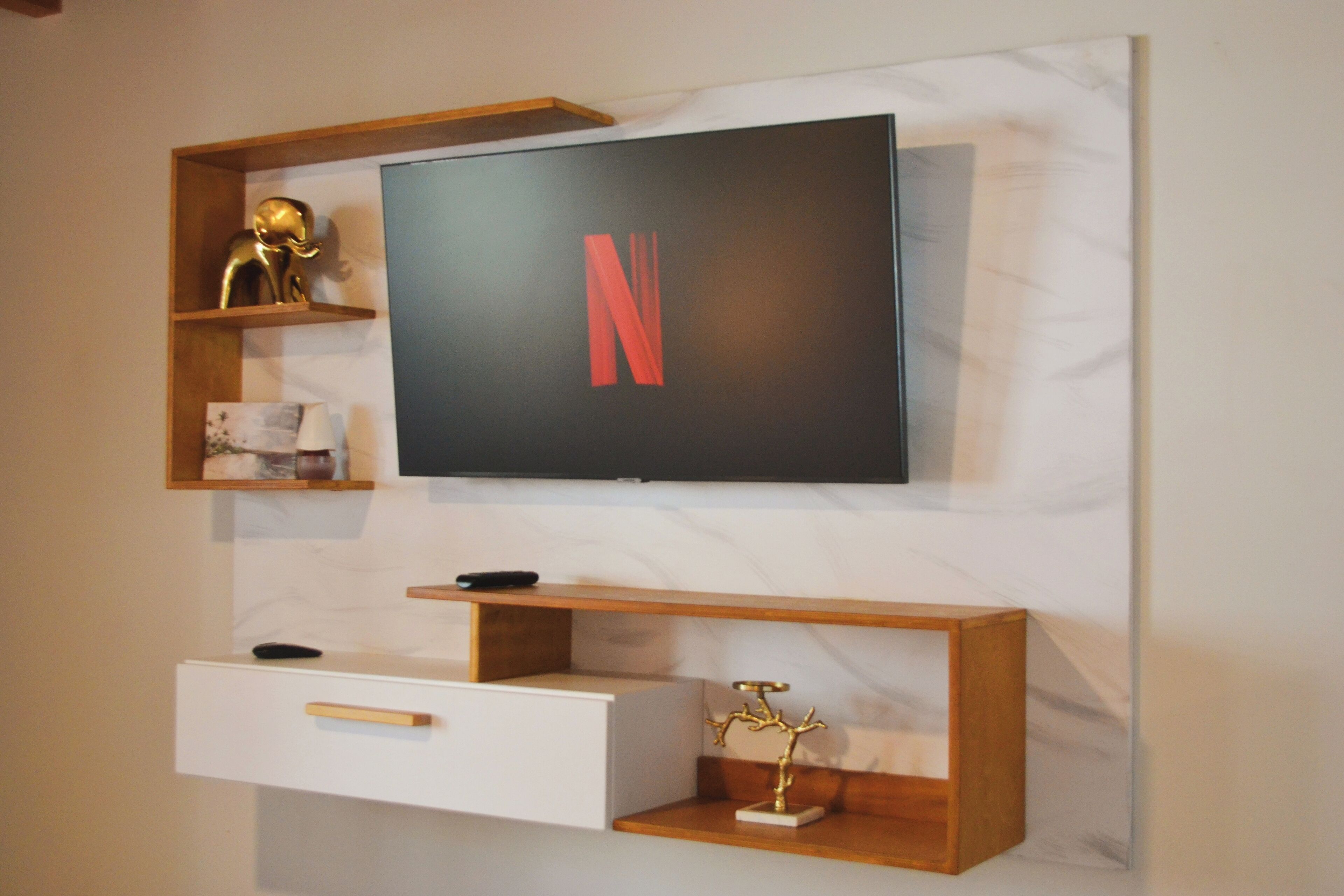 A wall-mounted TV with a Netflix logo, wooden shelves with decorative items, and a white drawer unit.