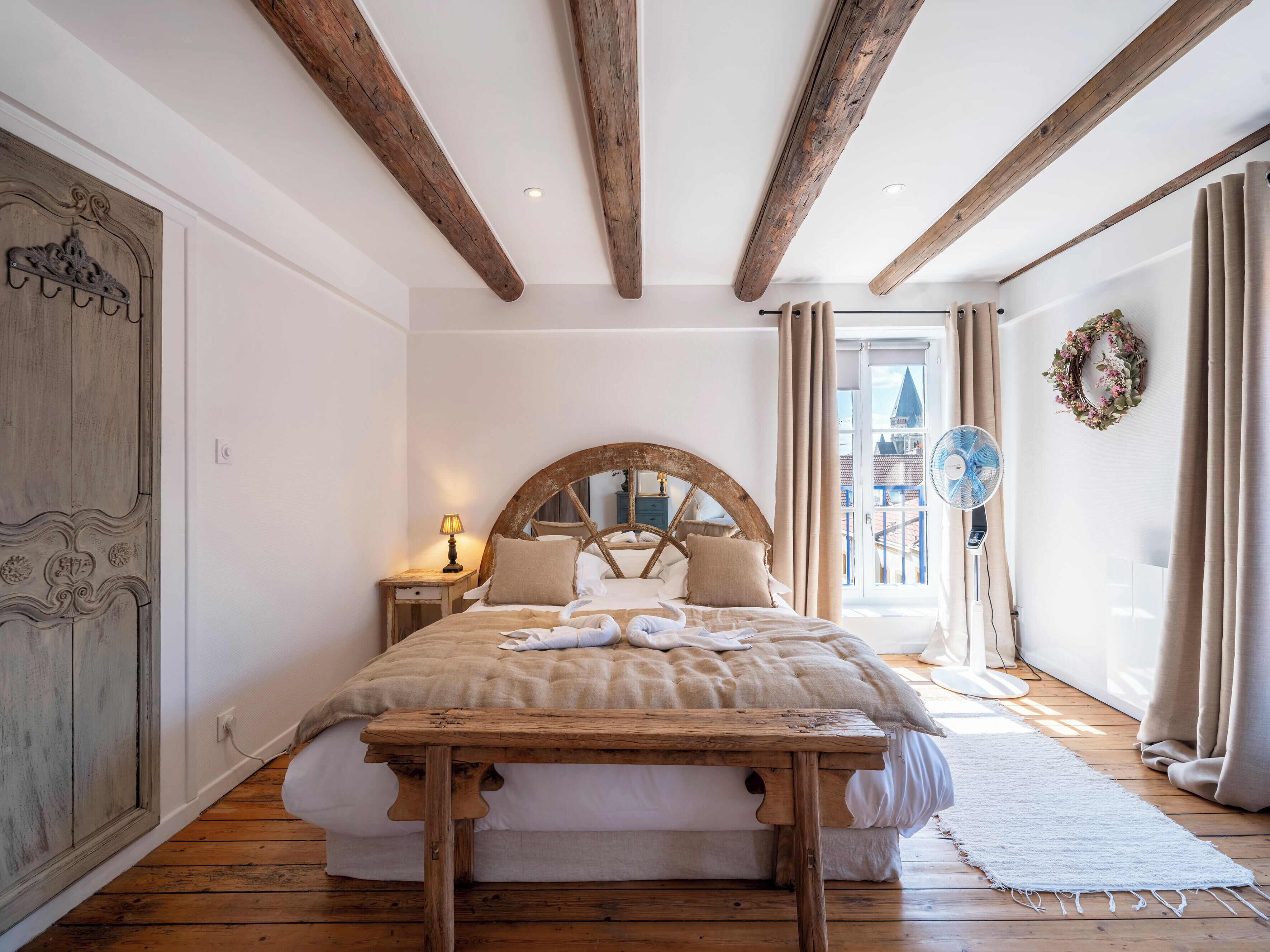 A bedroom with a large bed, wooden beams on the ceiling, a wooden table, and a view of the city through the window.