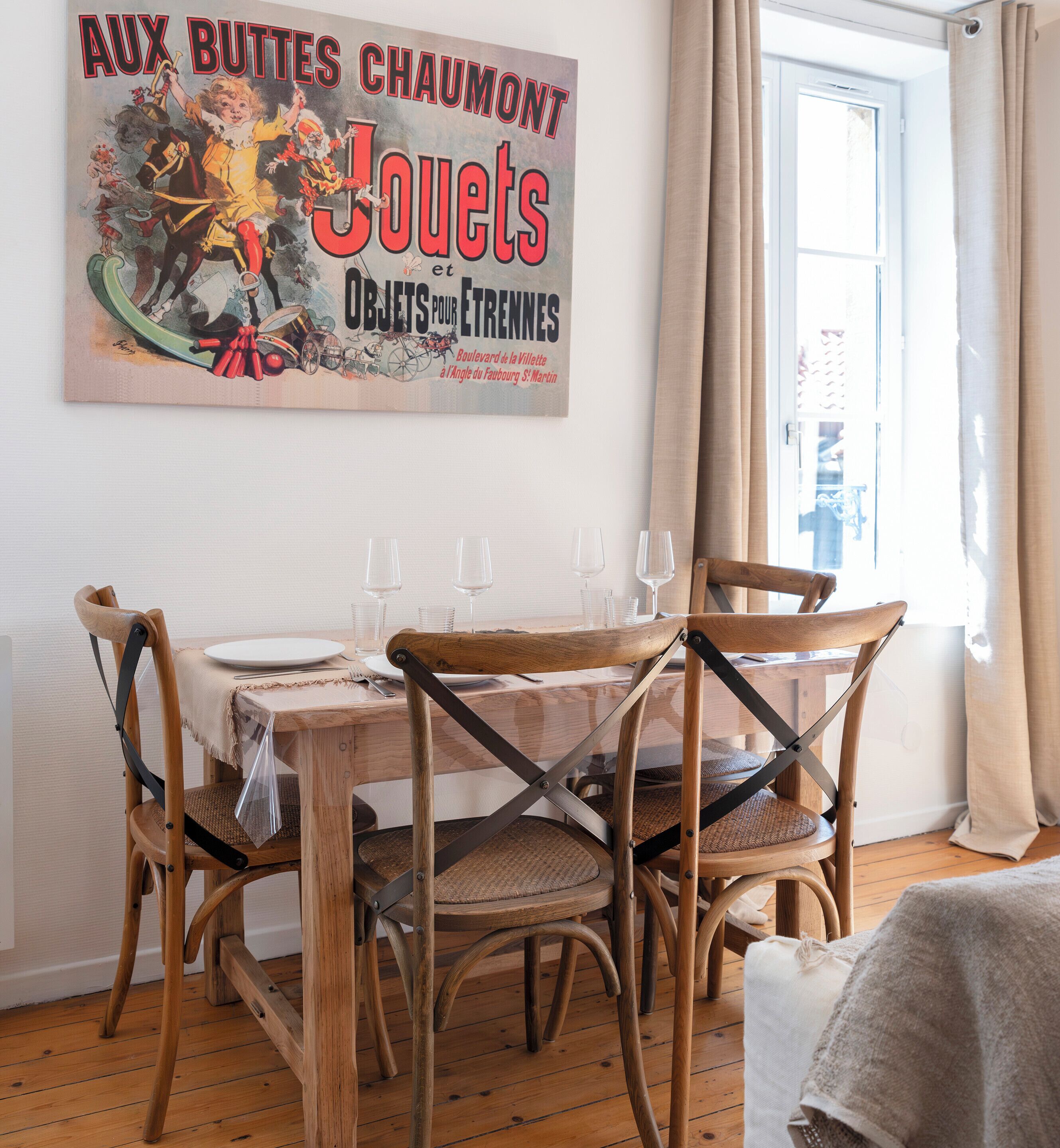 Un coin repas avec une table en bois dressée pour quatre personnes, entourée de chaises en bois à ossature métallique. Une affiche ancienne est accrochée au mur, au-dessus de la table.