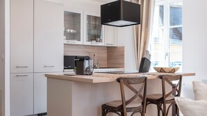 A modern kitchen with white cabinetry, a central island with bar stools, a black range hood, and a window with curtains. - Rock in Share - A la Fleur de Ly (Metz)