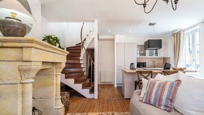 A modern living room with a wooden coffee table, a sofa with striped pillows, a stone fireplace, and a staircase leading to a kitchen area. - Rock in Share - A la Fleur de Ly (Metz)