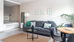 A modern living room with a grey sofa, a round wooden table, and a bed with a white headboard. There are three framed botanical prints on the wall, a floor lamp, and a potted plant. - Bright & Cozy, Renovated Studio in the Center (Lahti)