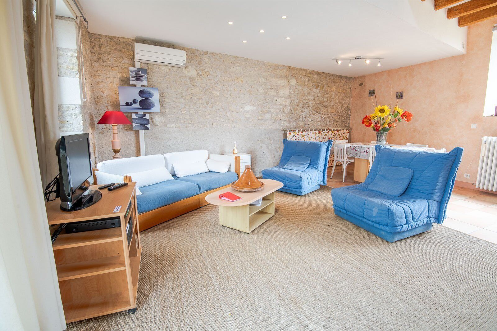 A cozy living room with a stone wall, a blue sofa, a blue armchair, a small table with a vase of flowers, and a TV on a wooden stand.