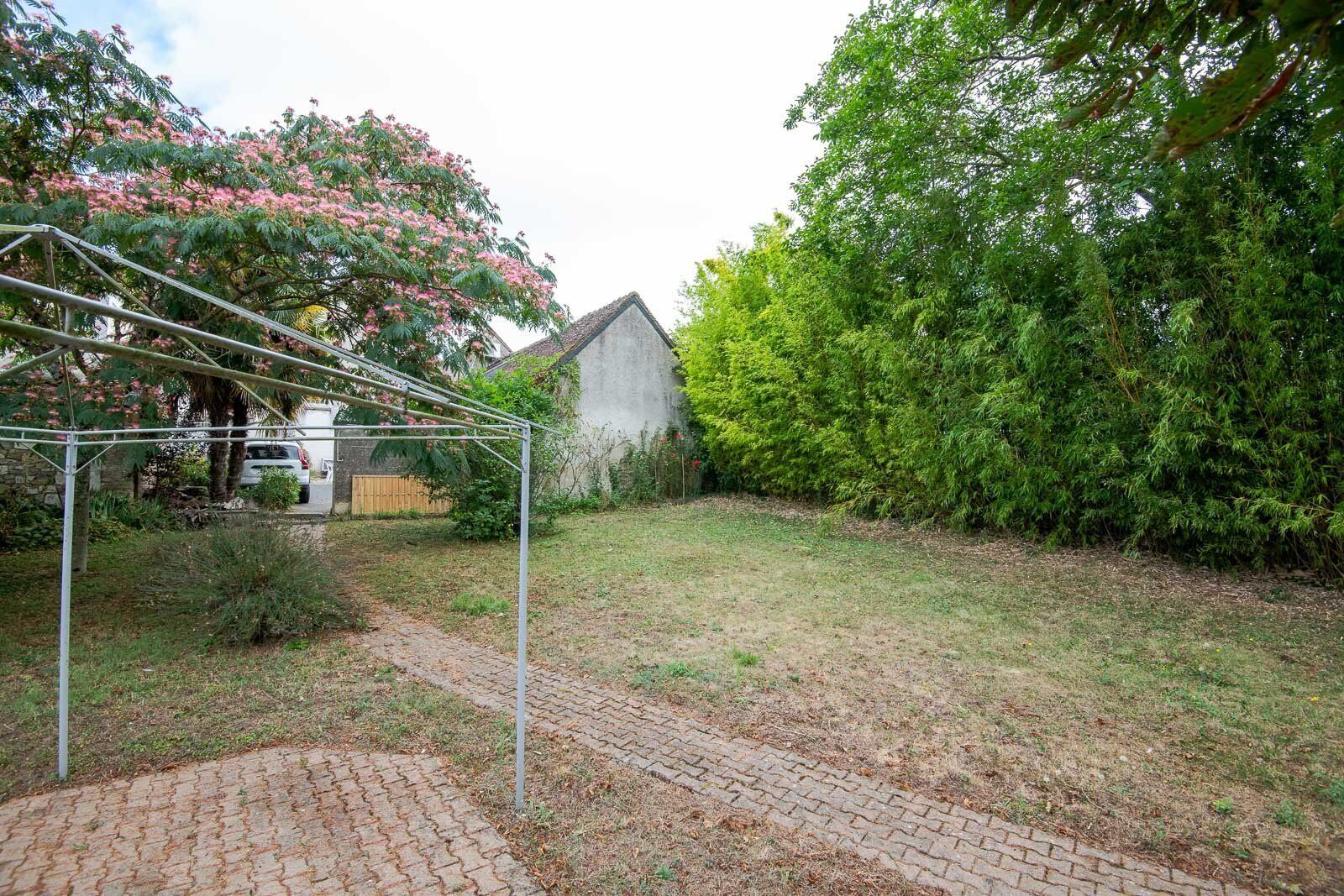 Un patio trasero con un camino de ladrillos, una pérgola de metal y diversas especies de árboles y arbustos.
