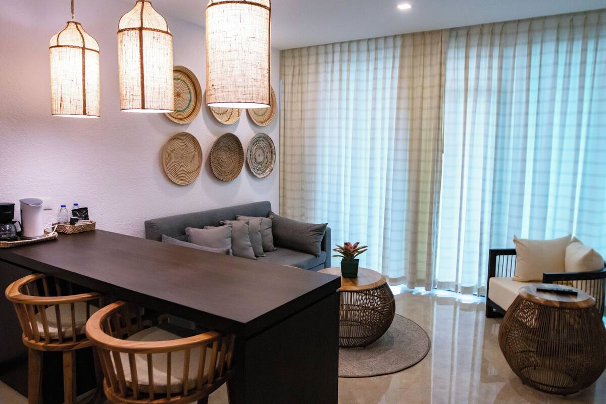 Una sala moderna con una mesa de comedor de madera oscura, dos sillas de madera, un sofá y una mesa de centro. Hay tres lámparas tejidas en el techo y un ventanal con cortinas.