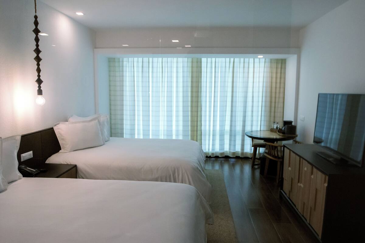 Una habitación de hotel con dos camas, una mesita, un televisor y una ventana con cortinas.