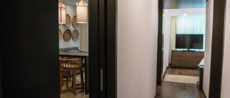 A hallway leading to a dining area with a wooden table and chairs, a wall-mounted light fixture, and a television in the background.