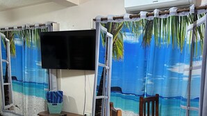 Ein Zimmer mit Fliesenboden, einem runden Holztisch, zwei Holzstühlen, einem an der Wand montierten Fernseher und einem Fenster mit Blick auf eine Strandlandschaft.