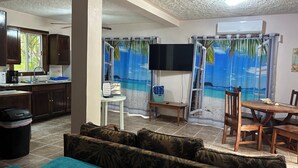 Ein Wohnzimmer mit Sofa, Esstisch und Fernseher. Es gibt eine Küchenzeile mit Schränken und einem Fenster.