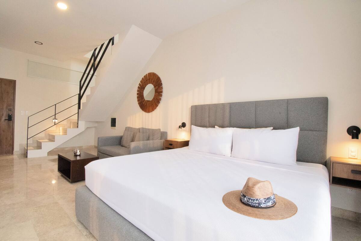 Une chambre d’hôtel moderne avec un grand lit, un canapé gris, une petite table basse, un escalier et un miroir fixé au mur.