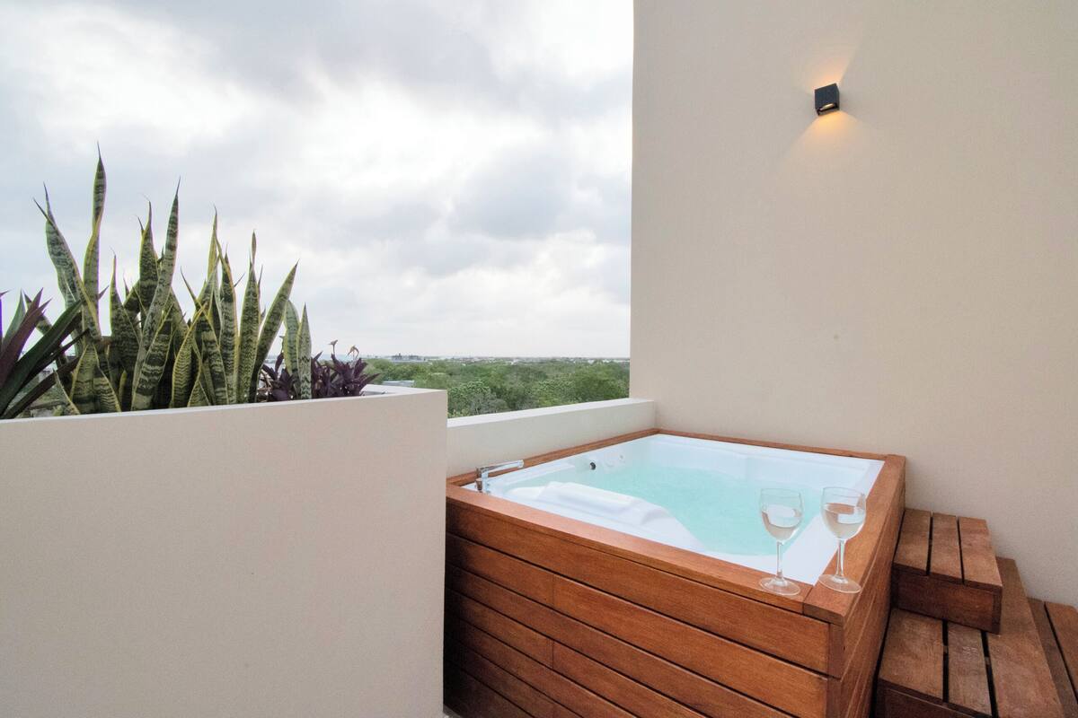 Un bain à remous en bois, avec deux verres de vin blanc, sur un balcon offrant une vue sur la verdure et un ciel nuageux.