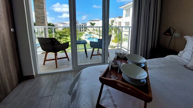A hotel room with a balcony, a bed, a tray with dishes, and a view of a pool and buildings. - Unique lake, Golf & Jacuzzi in Hard Rock Golf PC (Punta Cana)