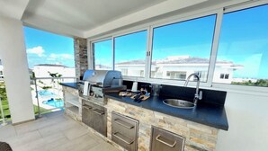 A modern outdoor kitchen with a stone backsplash, a built-in grill, and a large window overlooking a residential area. - Unique lake, Golf & Jacuzzi in Hard Rock Golf PC (Punta Cana)