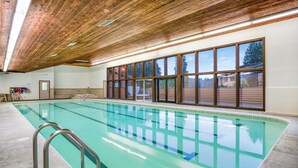 Piscina interna com água cristalina, vigas de madeira no teto e grandes janelas com vista para o exterior.