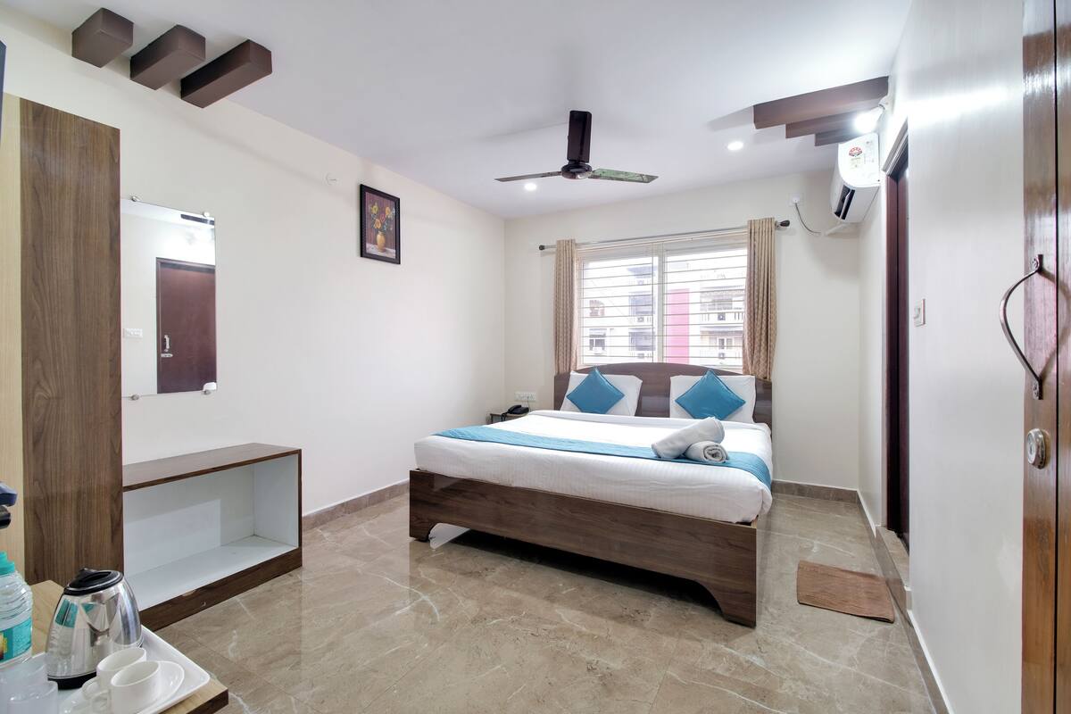 A hotel room with a bed, a bedside table, a mirror, a ceiling fan, and a window with curtains.