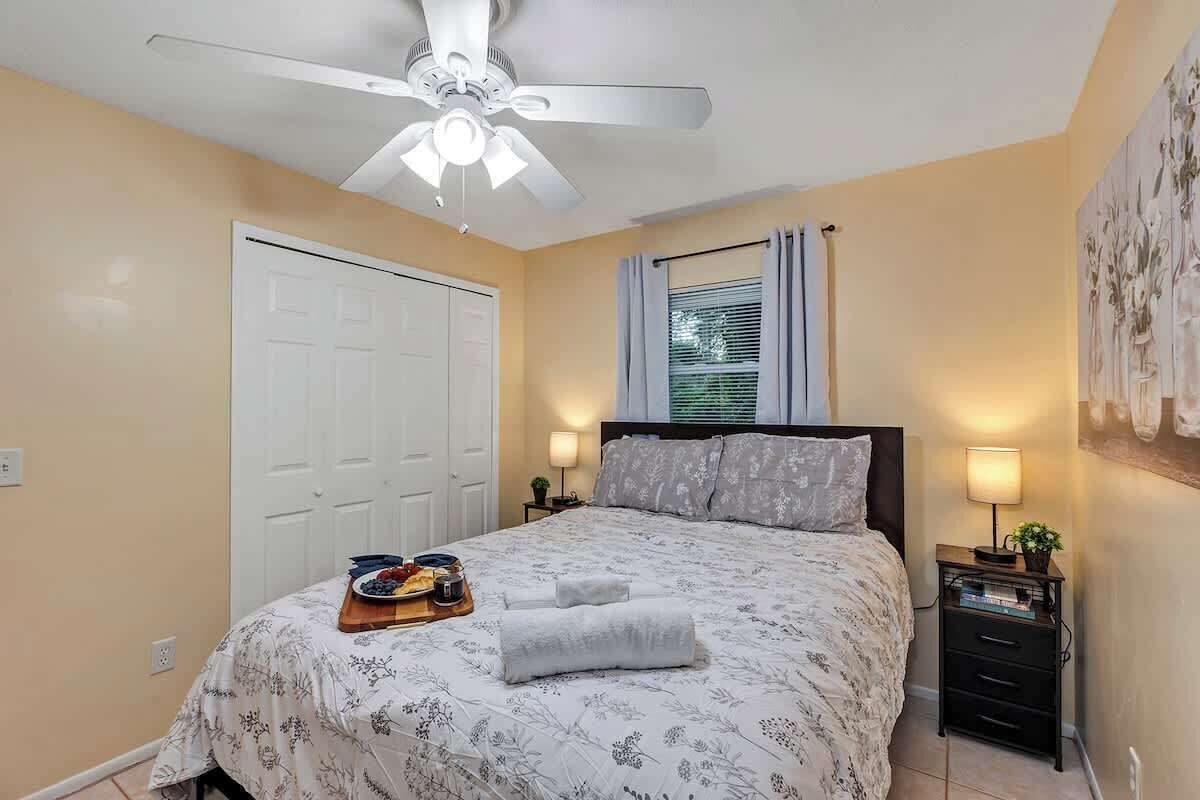 A bedroom with a bed, a ceiling fan, a window with curtains, a bedside table with a lamp, and a wall-mounted artwork.