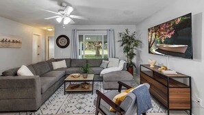 A modern living room with a sectional sofa, a coffee table, a TV, and a large window with curtains. - Cozy 4/3, Patio, King Bed, Bunk Room + Water Park! (Sanford)