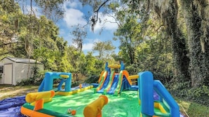 An inflatable play area with slides and climbing structures, surrounded by trees and a house. - Cozy 4/3, Patio, King Bed, Bunk Room + Water Park! (Sanford)