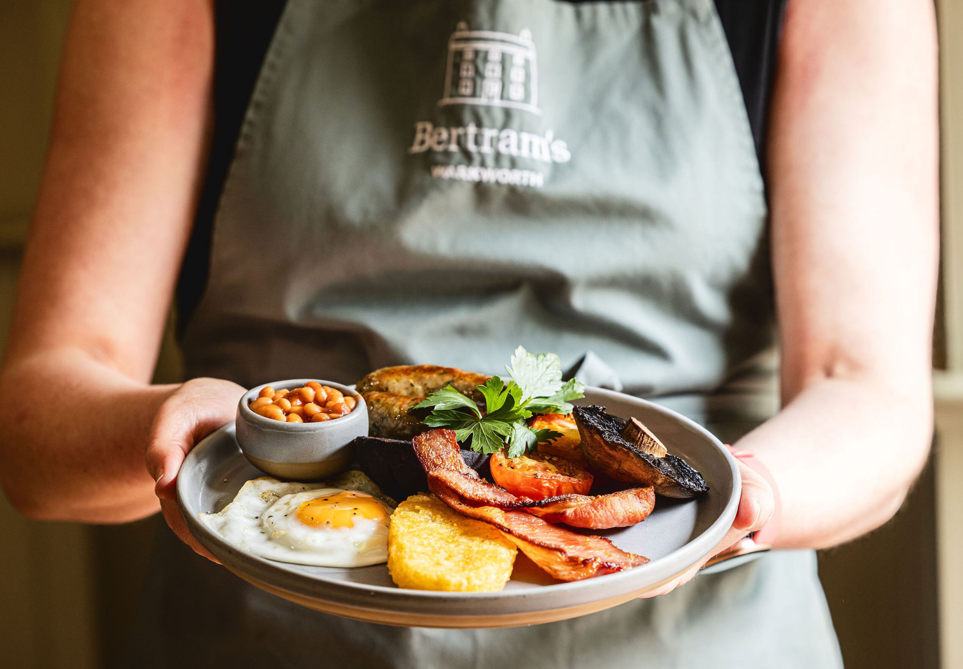 A person holding a plate with a breakfast meal including eggs, bacon, beans, mushrooms, and toast.