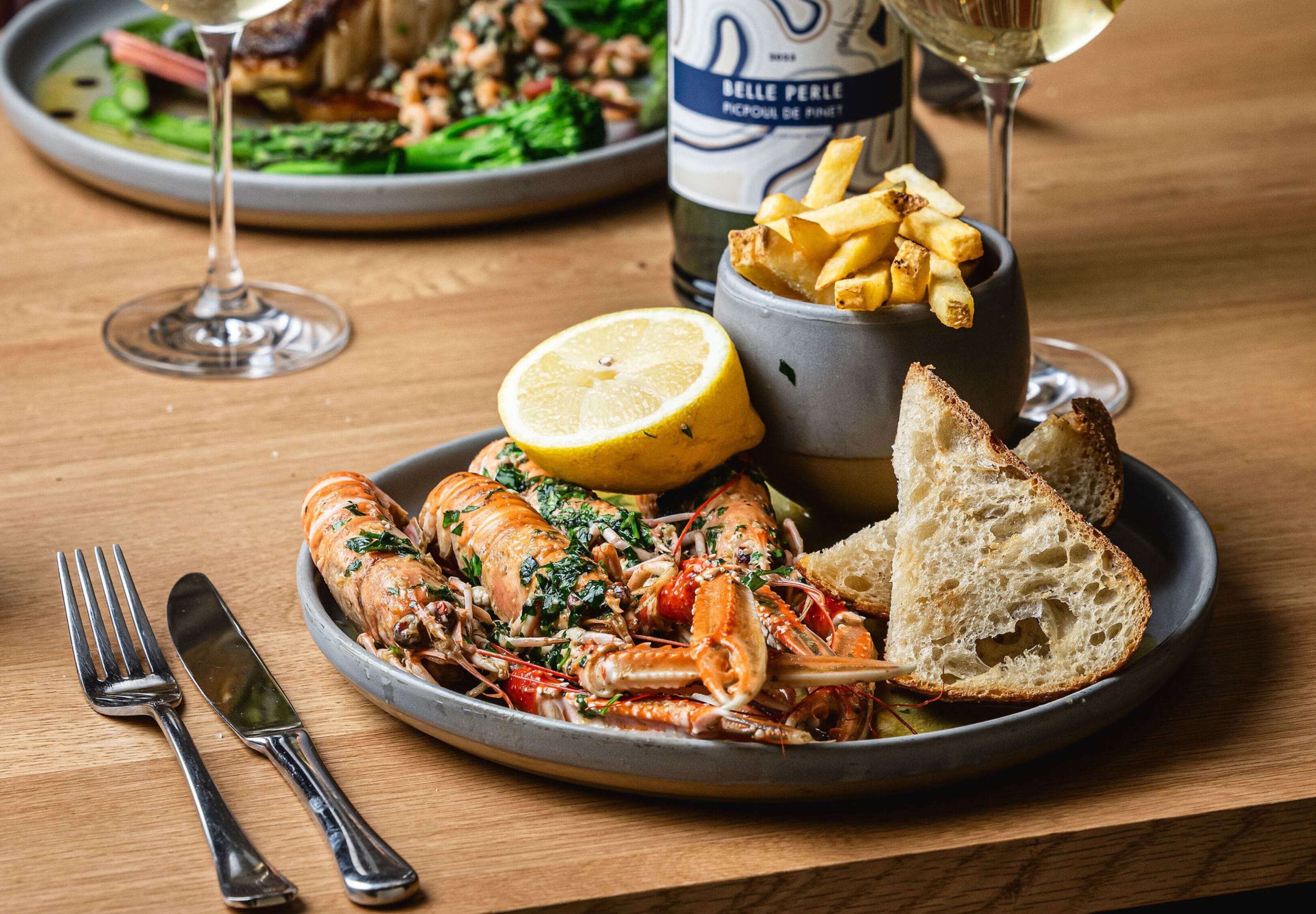 A plate of seafood, including lobster, accompanied by lemon, fries, and bread, with a bottle of Belle Perle wine and a glass of wine on the table.
