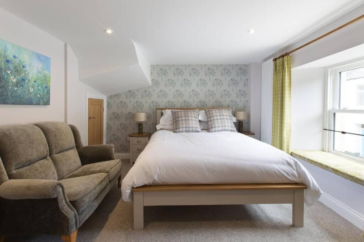 A bedroom with a bed, a sofa, a nightstand, a lamp, a window with curtains, and a painting on the wall.