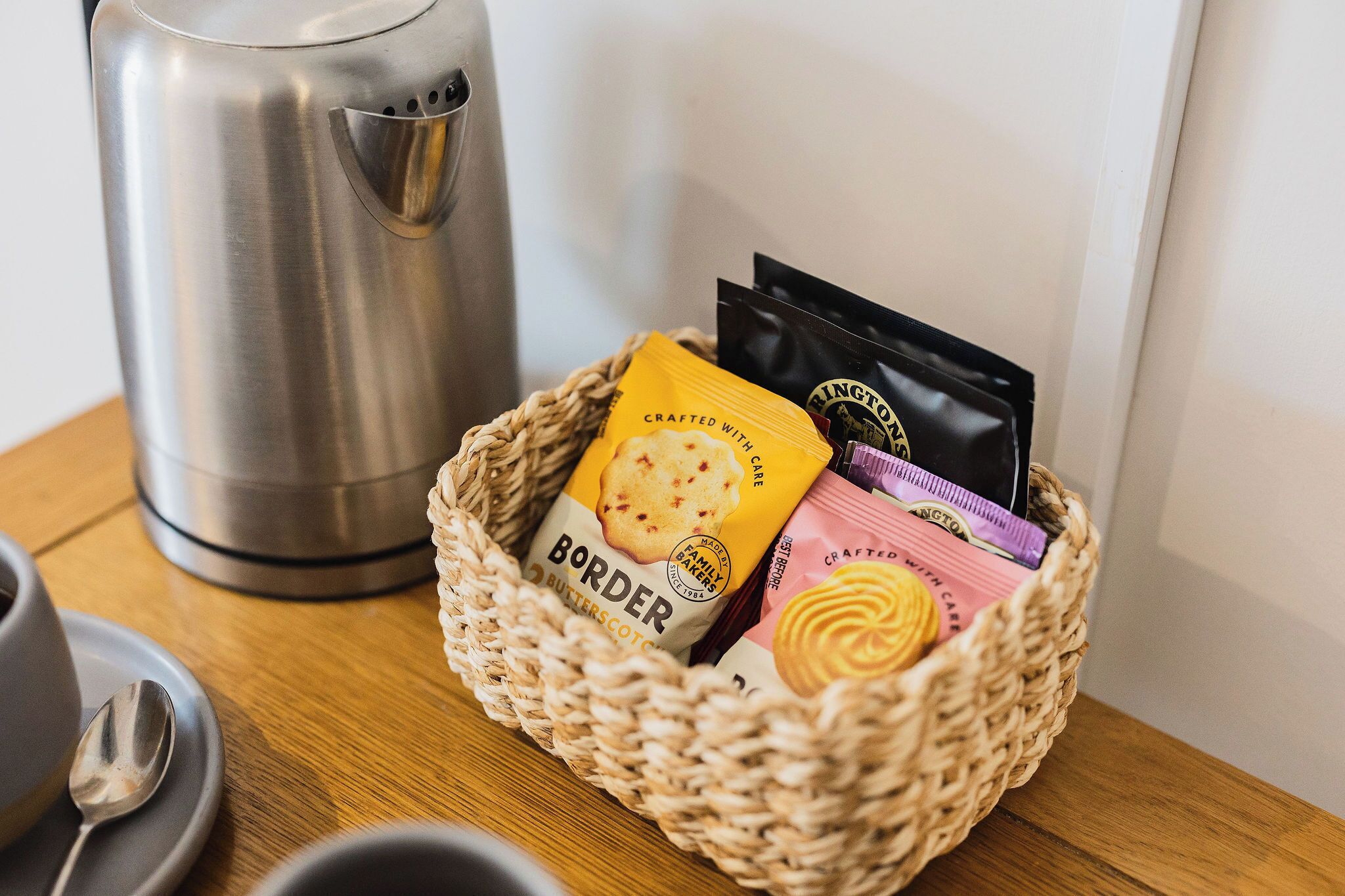 A woven basket containing a biscuit, a tea bag, and a coffee pod on a wooden table next to a stainless steel kettle.