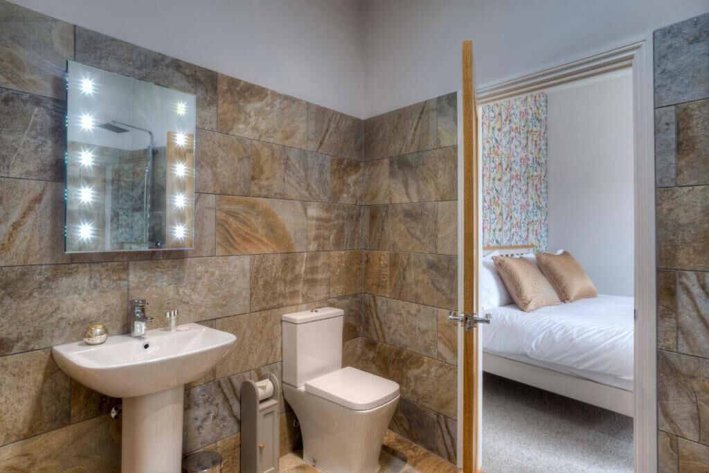 Baño con lavamanos, inodoro y espejo con luces. En la habitación contigua, hay una cama con almohadas.
