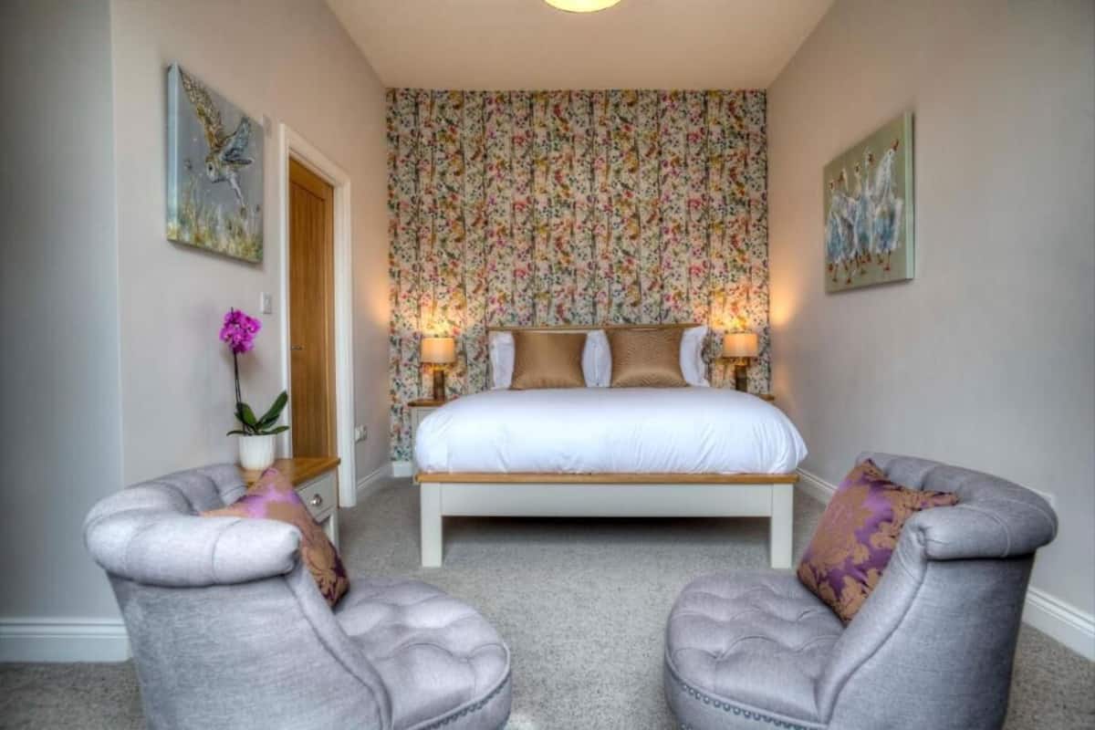 A bedroom with a bed, two armchairs, a nightstand, a potted plant, and floral wallpaper.