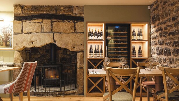 A cozy restaurant with a stone fireplace, wooden tables and chairs, and wine bottles on shelves.