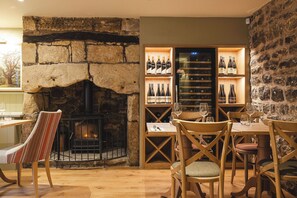 A cozy restaurant with a stone fireplace, wooden tables and chairs, and wine bottles on shelves. - Bertram's (Morpeth)