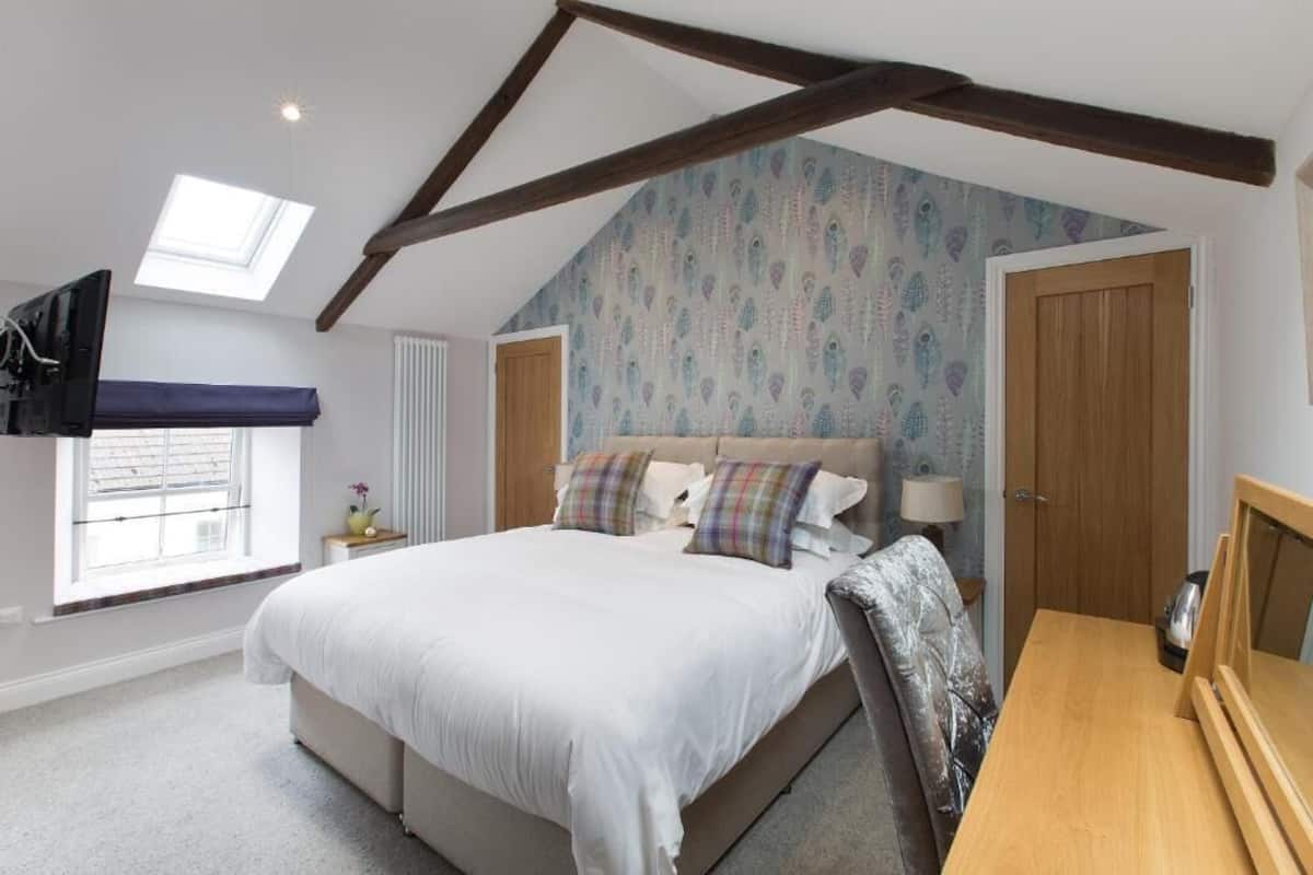 A bedroom with a sloped ceiling, a bed with multiple pillows, a window with blinds, a bedside table, and a wooden door.
