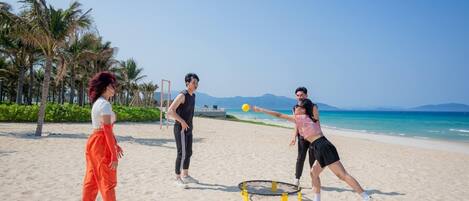 Three people playing with a ball on a sandy beach with palm trees and a clear sky in the background.