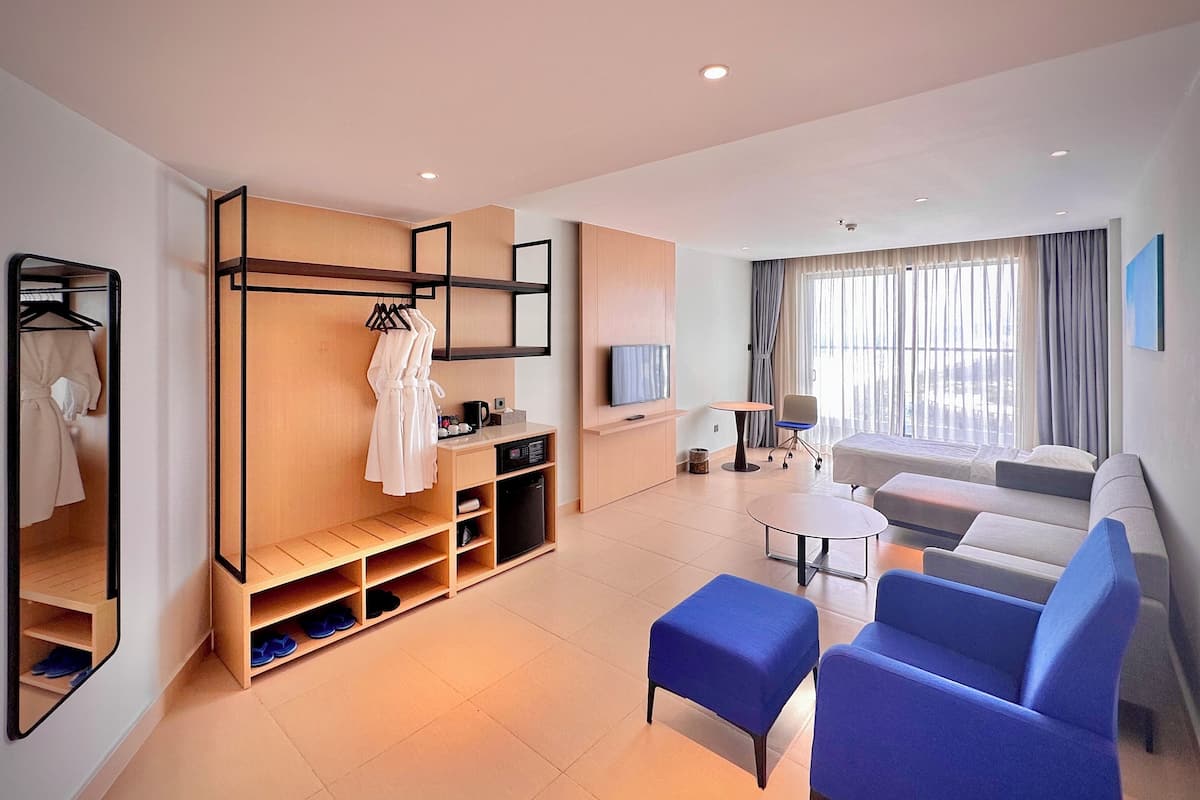 A modern hotel room with a sofa, ottoman, TV, and a seating area with a table.