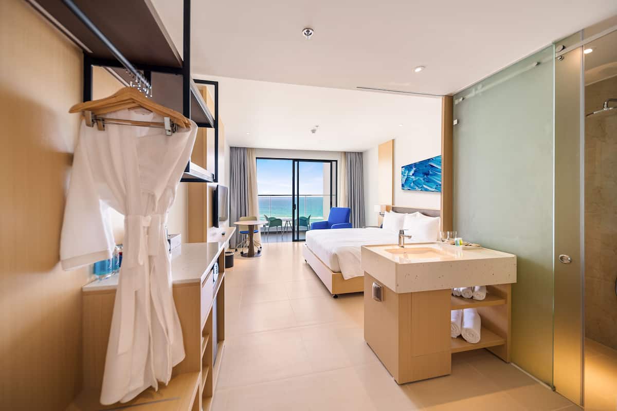 A modern hotel room with a large bed, a bathroom with a sink and mirror, a robe hanging on a rack, and a view of the sea through the window.