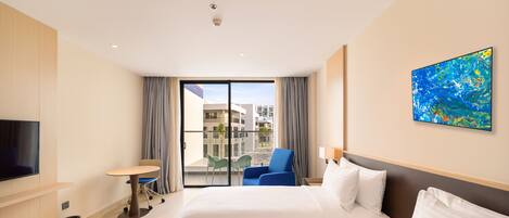 A hotel room with a bed, a desk, a chair, a TV, and a balcony with a view of buildings.