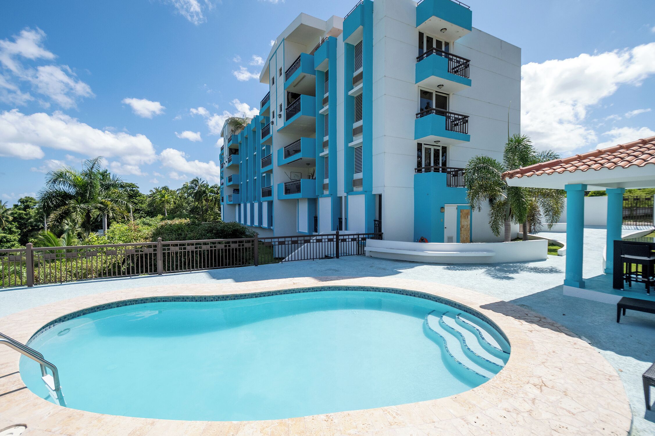 A swimming pool with a tiled deck, surrounded by a multi-story building with balconies, and palm trees in the background.