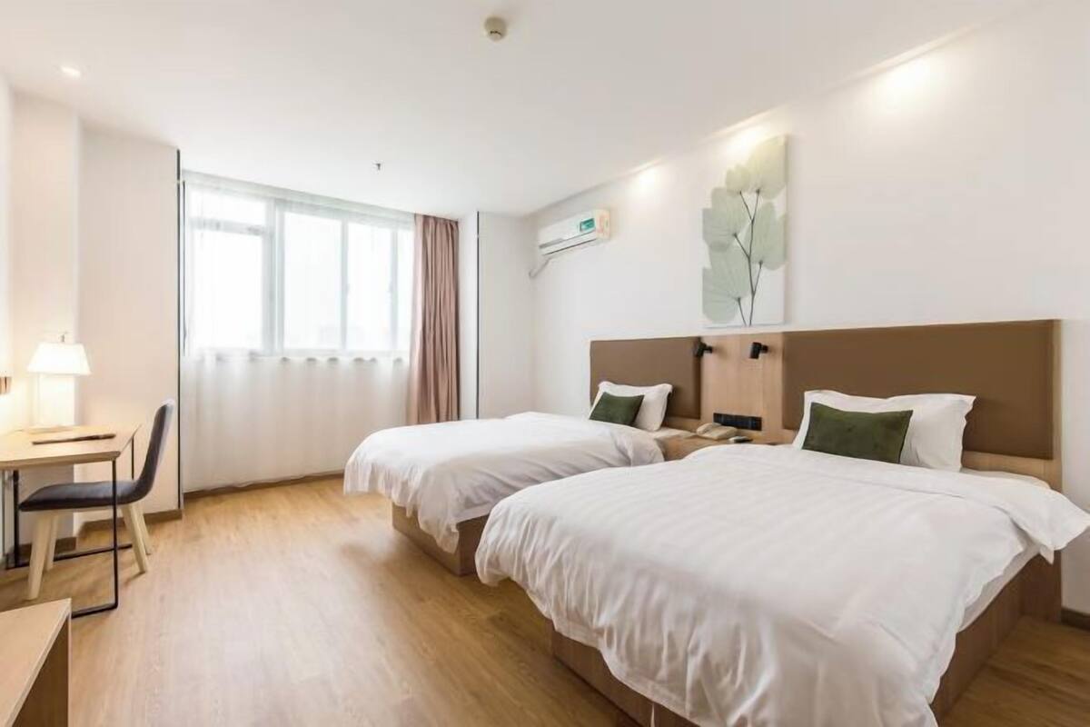 A hotel room with two beds, a desk, a chair, a window with curtains, and a wall-mounted air conditioner.