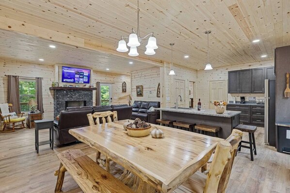 A spacious living room with a wooden dining table, a fireplace, and a large TV mounted on the wall.